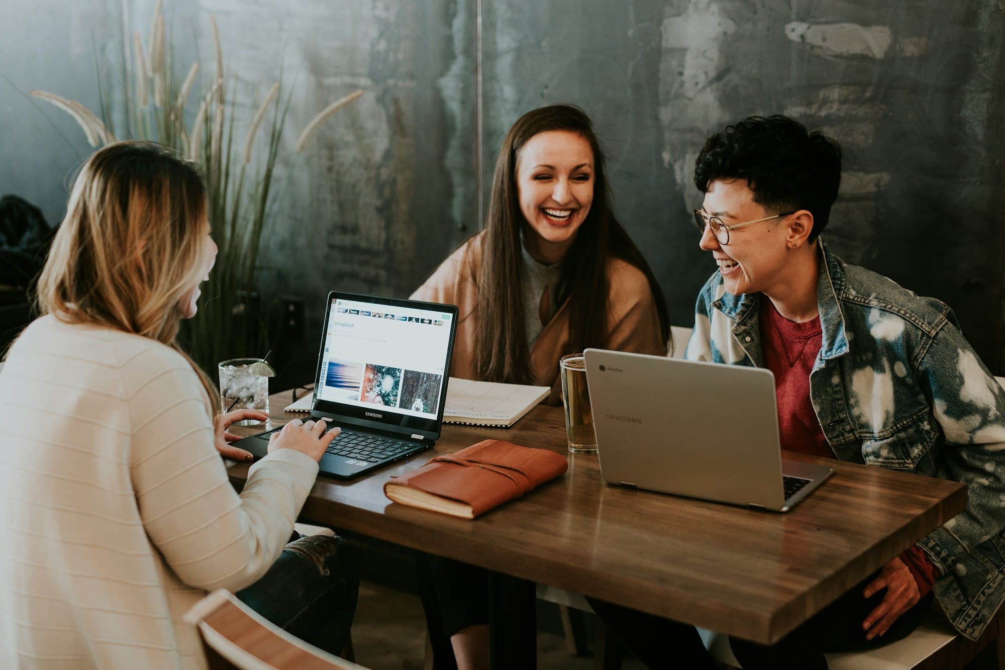 Three people laughing around a café table with laptops and a notebook